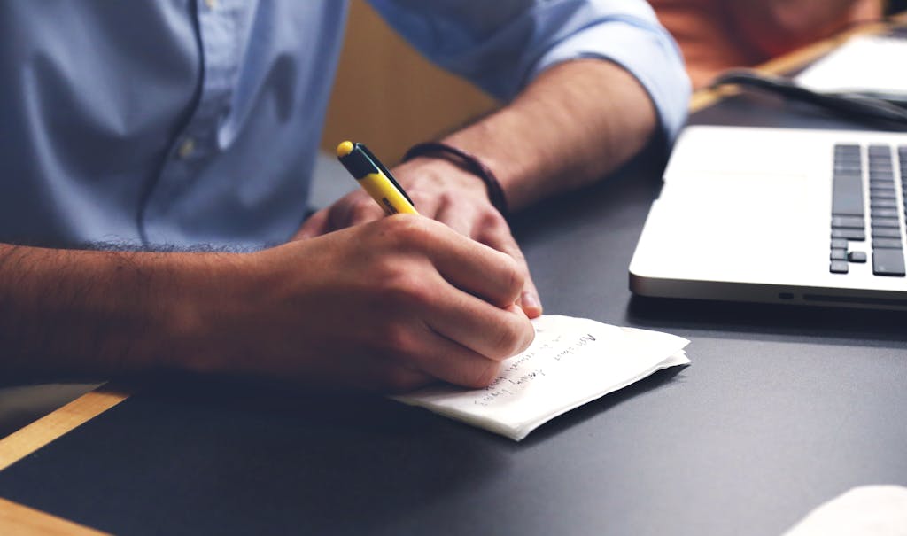 Close-up of a man writing notes with a pen next to a laptop on a desk.