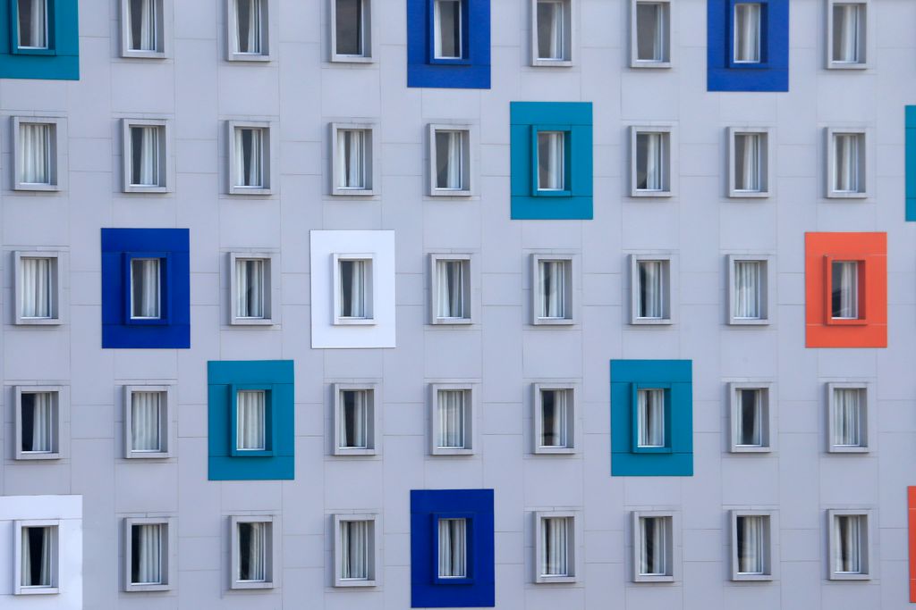 White building facade covered in rows of small windows, some framed in blue, teal, orange and white, suggesting dozens of separate sites under one roof