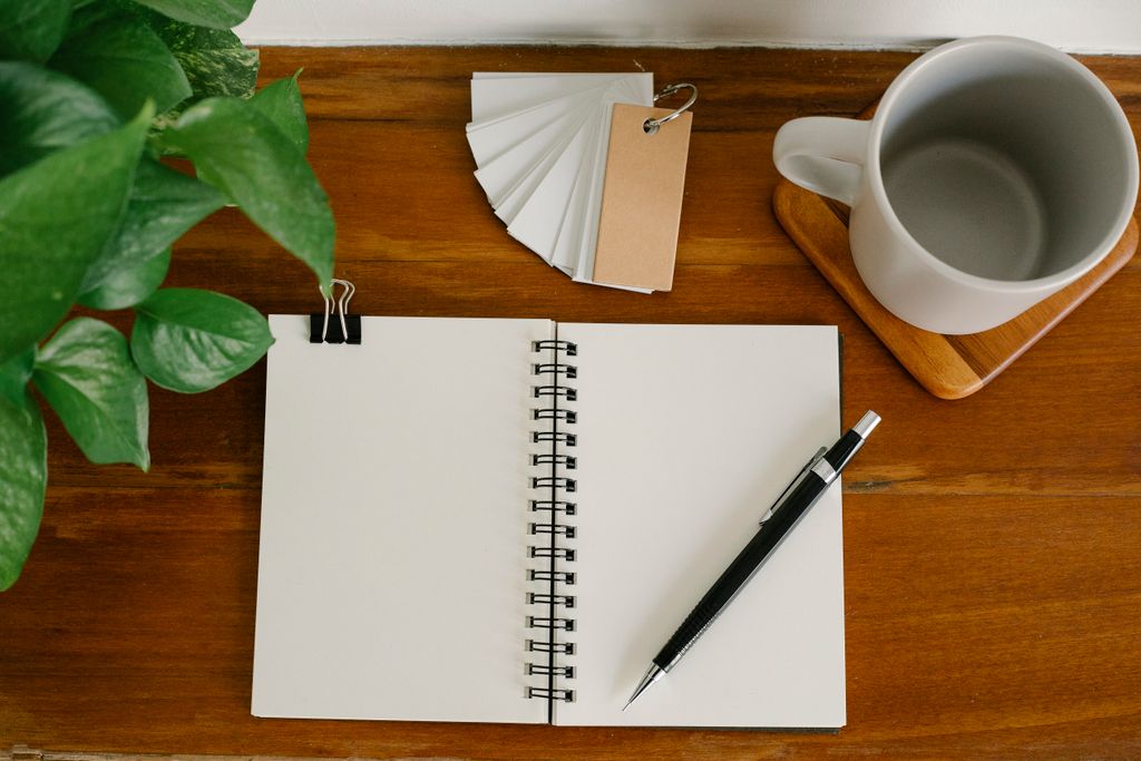 Open blank spiral notebook with a mechanical pencil on a wooden desk, beside a white mug, a potted plant and a fan of blank cards